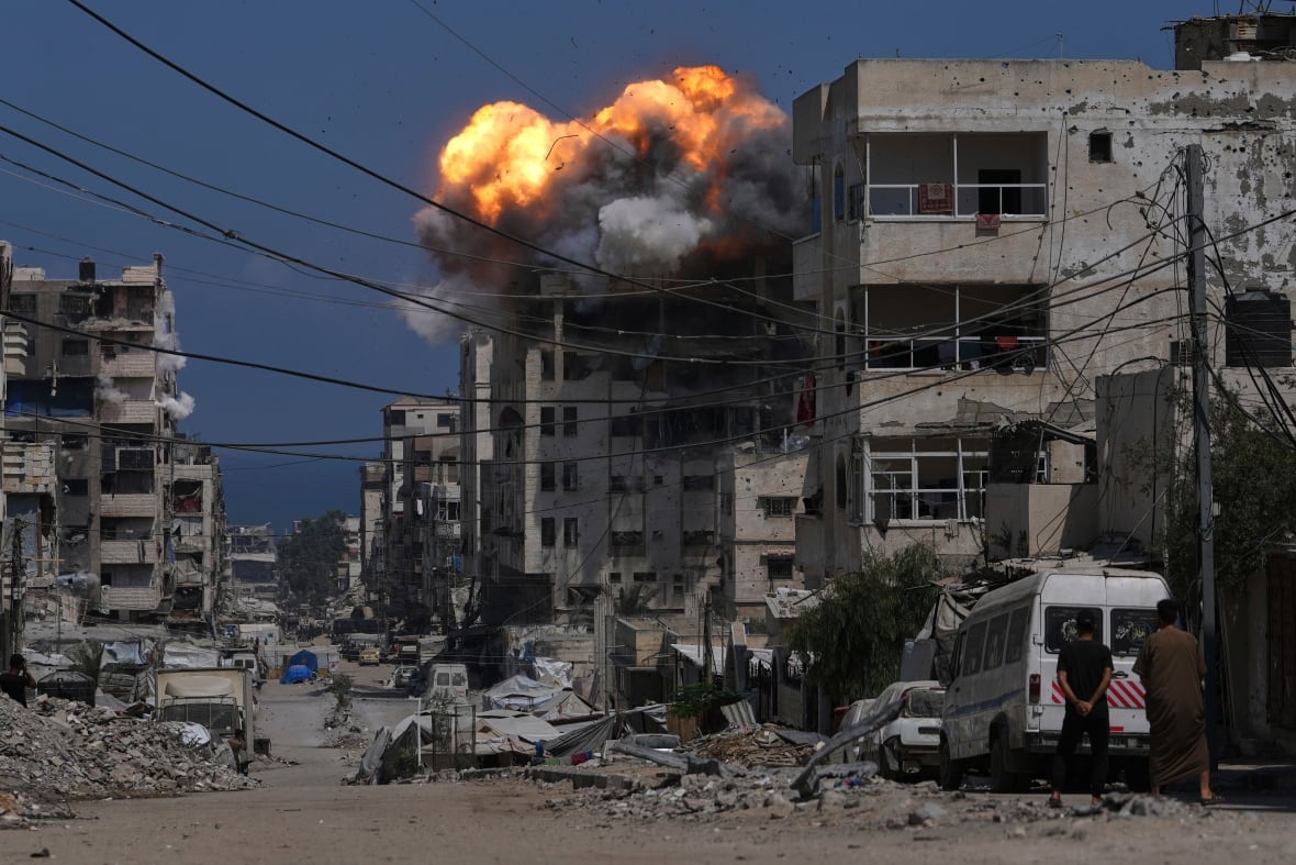 The top of a building is englufed in flames in a war torn neighbourhood.