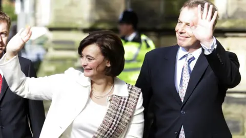 PA Media Cherie and Tony Blair wave and smile. She has short dark hair and is wearing a cream suit and top with a Burberry-pattern scarf hung over her left shoulder. He is wearing a dark suit and a silver tie with a red pattern