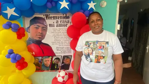 Gustavo Ocando Edwuar Hernández Herrera mother smiles at the camera. She is wearing a white T-shirt with photos of her son stamped on it. On the wall hangs a posted with another photo of her son and the message "Welcome home, my love". Balloons in the colours of the Venezuelan flag surround the poster