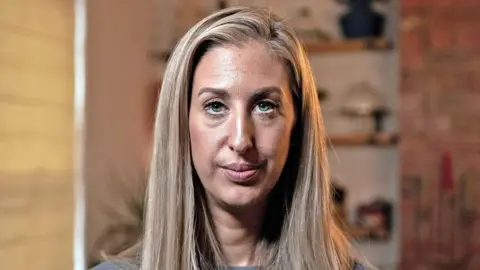 MARTIN MCQUADE / BBC Portrait photo of Fiona Winser-Ramm. She has long, straight blonde hair with a side parting. She has blue eyes and is wearing a grey ribbed jumper. She is pictured in a living room, with shelves and a blind visible behind her. 