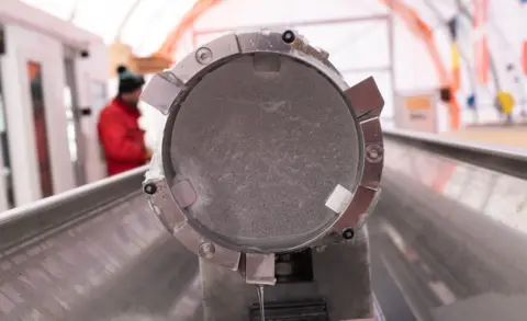 PNRA/IPEV The end of an ice core suspended in a metal tube, inside a metal gulley. It is inside a large tent in Antarctica where the ice cores are being kept. On the left-hand side, there is a person wearing a bright red jacket and a bobble hat.