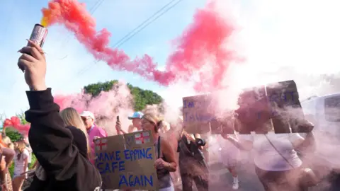 PA Media Red smoke pours out of a flare held up by a person who is part of a larger group marching along a road. Some are holding cardboard placards.