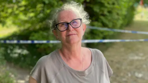 Sam Read/BBC A woman wearing glasses and a grey top is stood in front of some bushes. Between her and the bush is blue and white police tape.