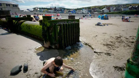 Getty Images A boy plays in a stream in front of a discharge pipe on a sunny beach in Wales, with sand and sunbathers in the background. 