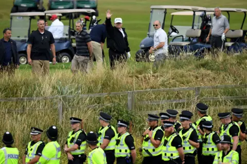 Getty Images Police secure the area as U.S. President Donald Trump, wearing a white hat with Trump and USA displayed on it, waves while playing golf at Trump Turnberry Luxury Collection Resort during the President's first official visit to the United Kingdom.