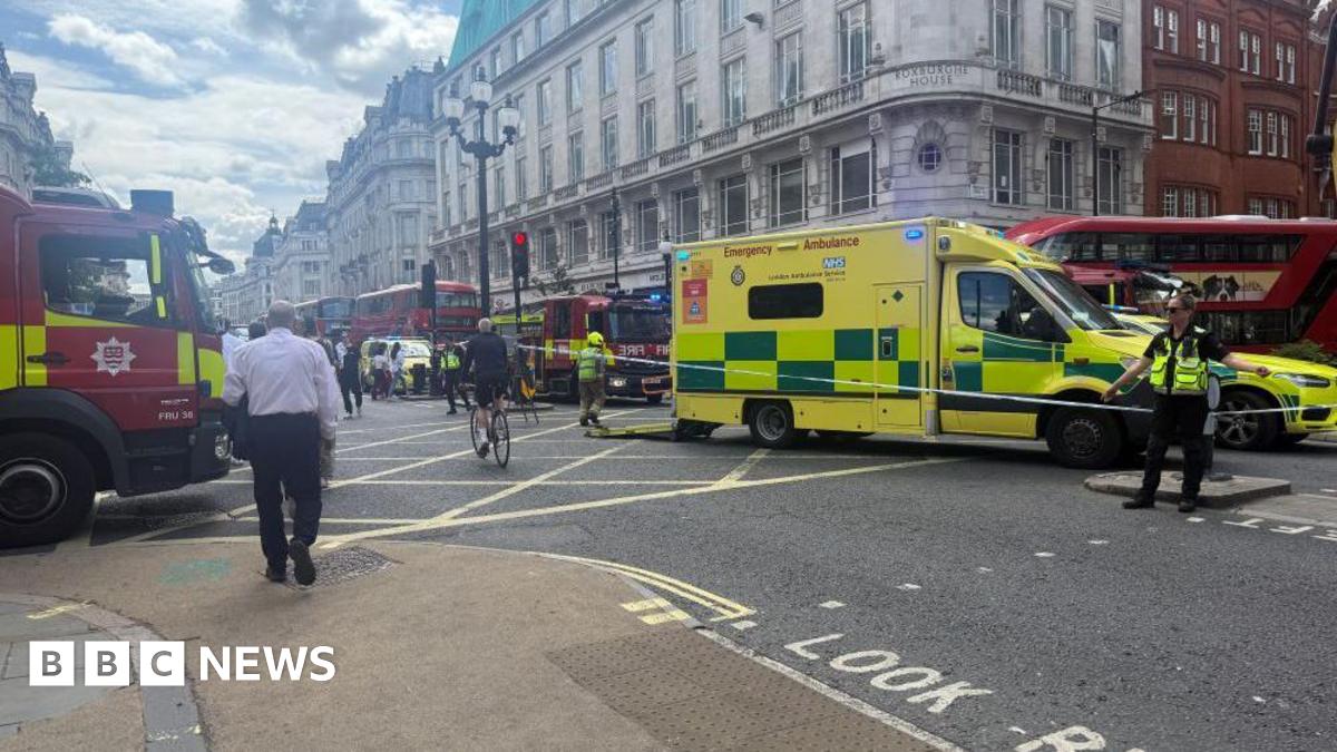 A number of emergency vehicles parked on a road junction. There is a yellow ambulance parked side on in the middle right, with police tape running in front of it and an officer in high-vis directing people away from the scene. A fire engine is parked on the left, with the front of the truck visible. A red double-decker bus is stationary behind the ambulance. Behind the ambulance, there is another red fire engine and a queue of two red double-decker buses. There are tall buildings on all sides.