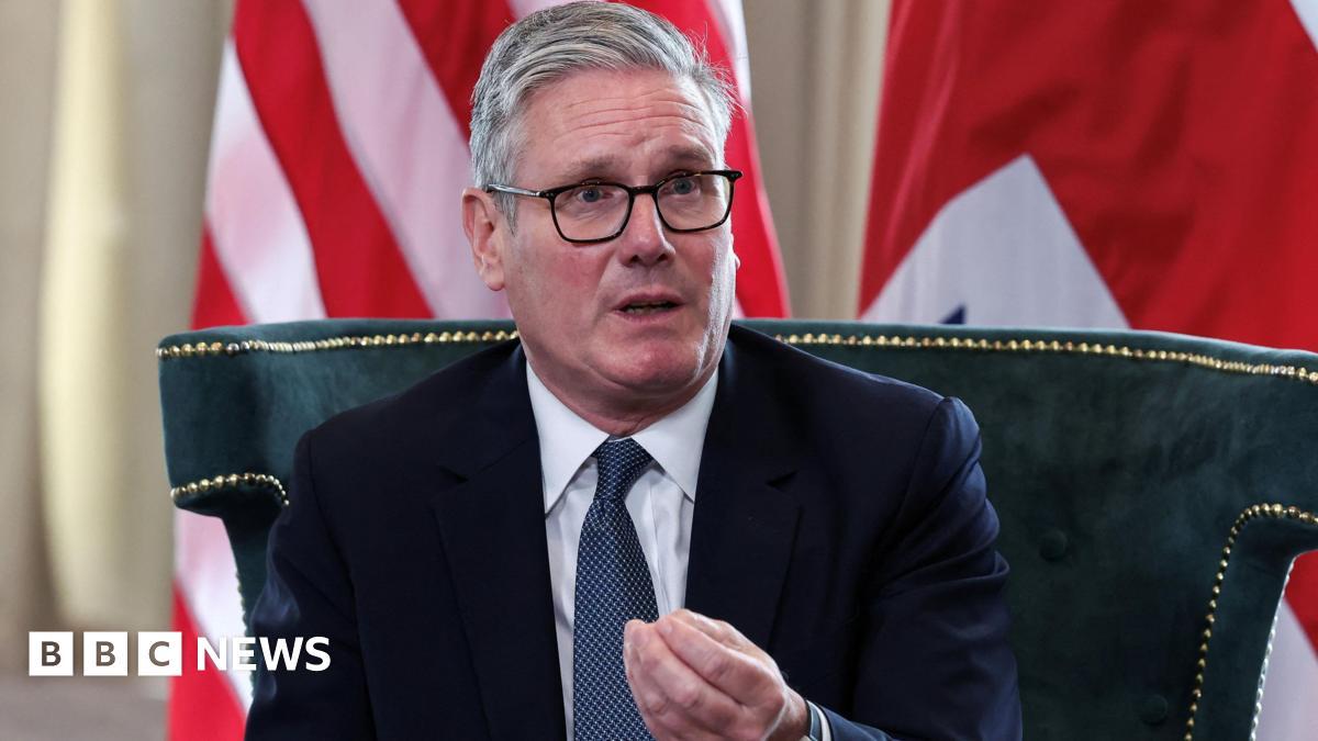 Sir Keir Starmer, sits on a green upholstered chair with gold trim, wearing a dark suit, white shirt, and blue patterned tie. He gestures with his right hand.