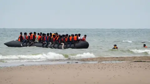 PA Media Near the shore at Wimereux, France, a group of people believed to be migrants, wearing orange life jackets, board an overcrowded inflatable boat. Some are already seated while others are still in the water trying to climb aboard. Waves crash onto the beach in the background.