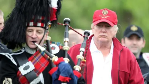 Getty Images A bagpiper in full military Highland dress with s bearskin hat is playing in the foreground on the left. Behind him and to the right is Donald Trump, in red Trump Golf baseball cap, white shirt and red fleece jacket. He is staring intently at the piper, looking very serious. There are others in golf gear in the background, just out of focus and slightly obscured by the President. 
