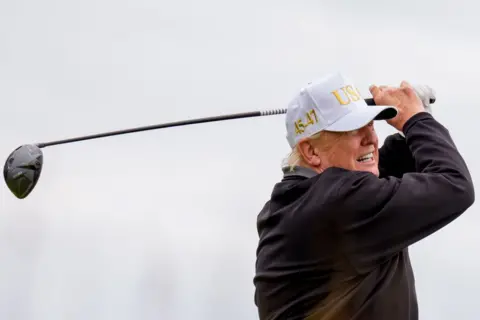 Getty Images President Donald Trump tees off at a new 18-hole course at Trump International Golf Links in Balmedie, near Aberdeen. He is wearing a white USA baseball cap and is at the end of his golf swing with the driver held aloft behind his head