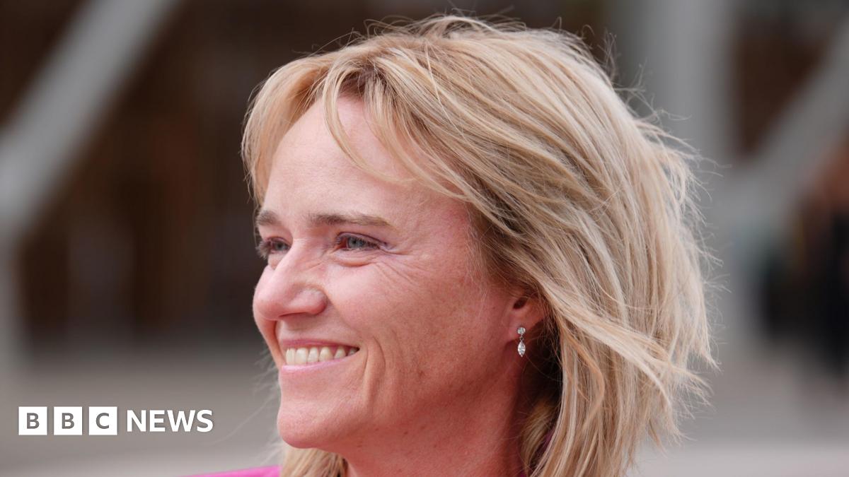 Sandie Peggie - a woman with blonde hair to her shoulders and a pink jacket smiles while standing outside