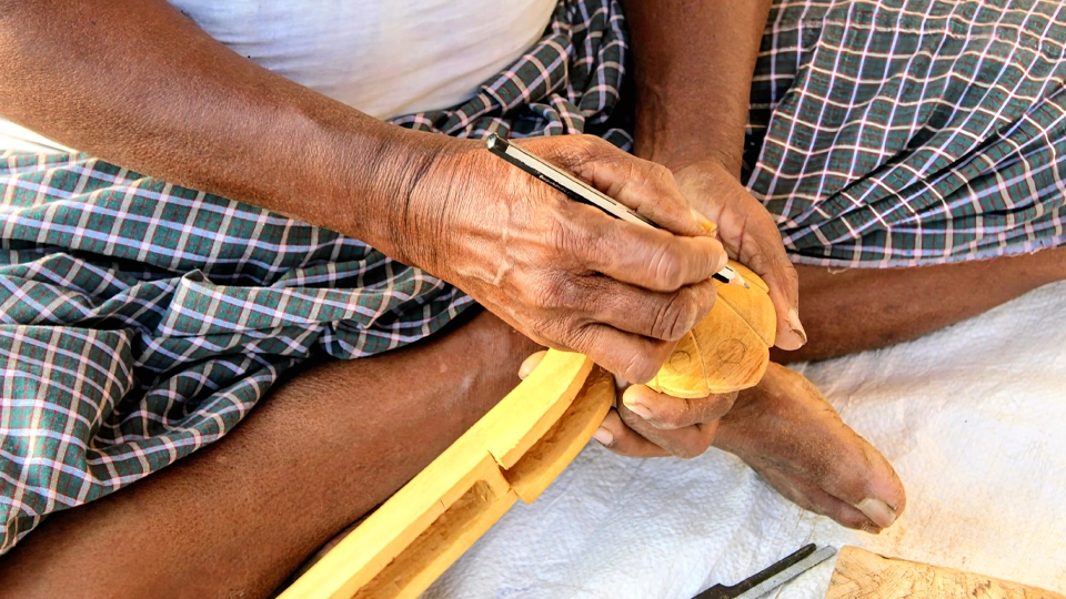 Artisan hand-carving a miniature Bobbili veena with a pencil on unfinished wood.