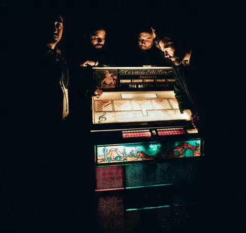 Cole Silberman Four band members standing in semi-darkness around an old-fashioned lit-up jukebox with "The Cosmic Selector" written in decorative lettering on the top
