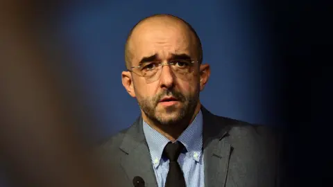 Getty Images A man is looking towards the camera. He wears glasses and has a grey suit on. Something is partially obscuring the front of the photo. 