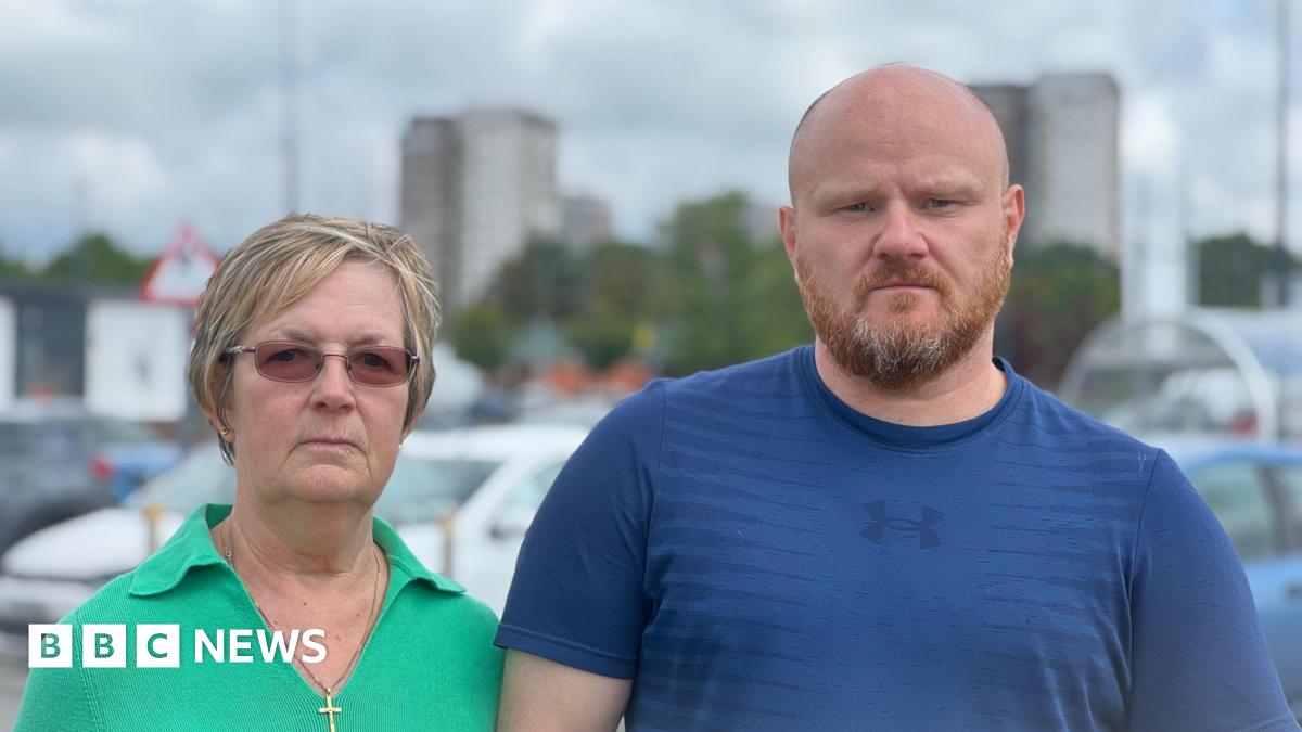 Elaine Shaw, who is wearing a green blouse and glasses and has ash-blonde hair, is standing next to Michael Jones, who is bald, has a ginger-grey beard and is wearing a blue t-shirt. They are standing in front of parked cars with Beech Rise and Willow Rise in the background behind them.