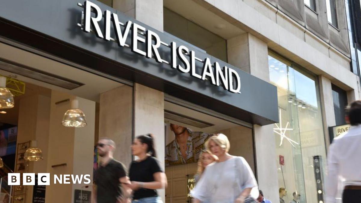 Shoppers walk past a River Island store on Oxford Street in London. It's a sunny day. Two friends are walking side by side and a woman with short blonde hair is walking.