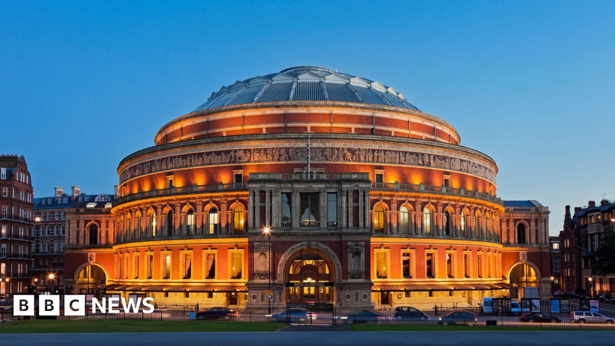 Exterior view of the Royal Albert Hall in London at dusk, with its red brick facade illuminated by golden lighting against a clear blue sky.