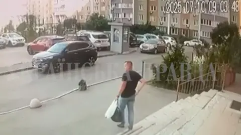 Reuters A man in jeans and a dark t-shirt holding a plastic bag and a holdall in one hand walks out of a building towards a car park in Kyiv.