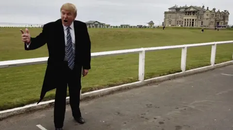 Getty Images Donald Trump in black coat and suit with white shirt and blue and white striped tie, standing on the road by the side of the 18th fairway to the Old Course, St Andrews. He is pointing and shouting, in a pose associated with his TV programme The Apprentice. The Royal and Ancient building is behind him and the sky is grey.
