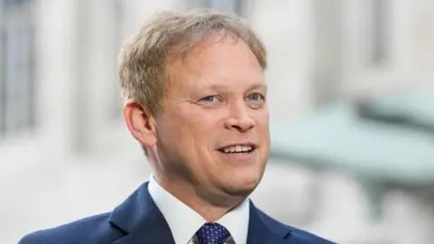 PA Media Grant Shapps has fair hair and is wearing a navy jacket over a white shirt and blue and pink tie. He is smiling with his mouth open.
