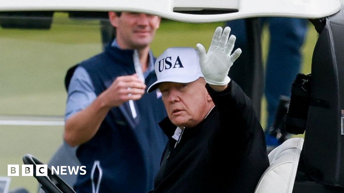 Donald Trump, wearing a blue jumper and trousers and a white USA cap, takes a golf swing on the fairway as other group members watch next to golf buggies
