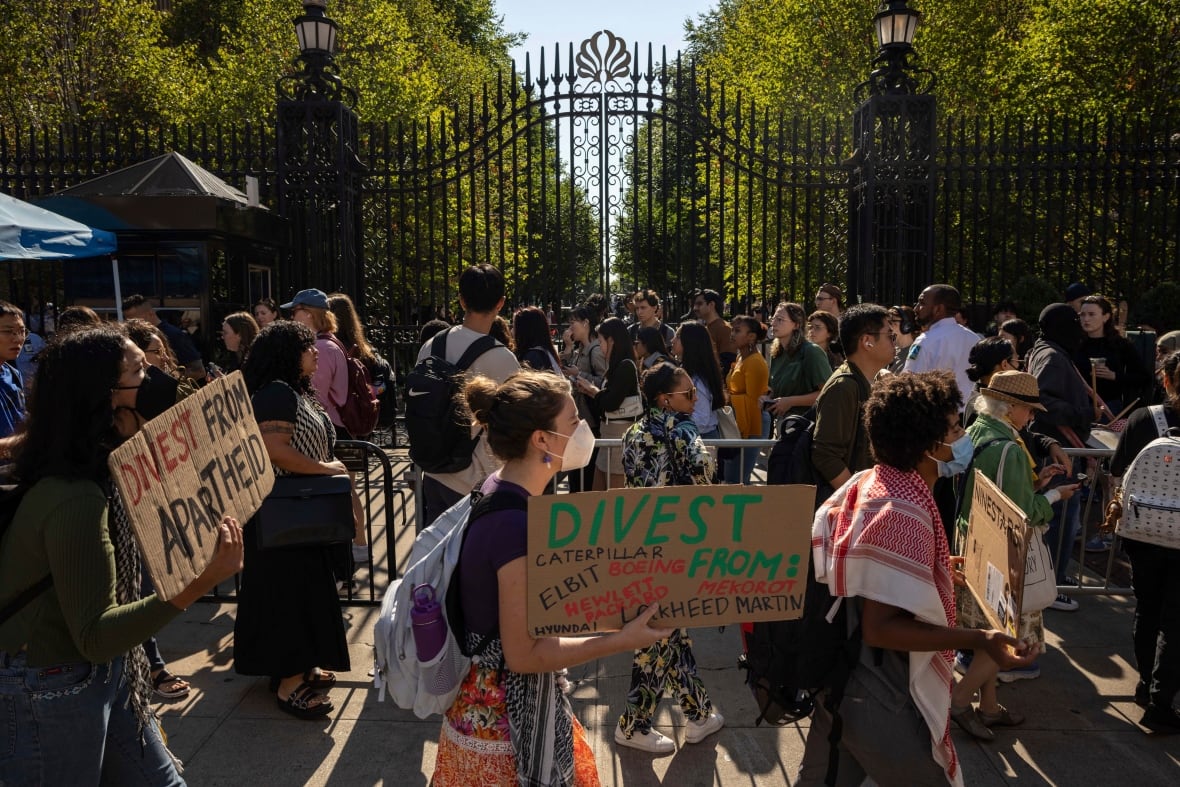 Young people walking past a wrought iron gate with signs and placards