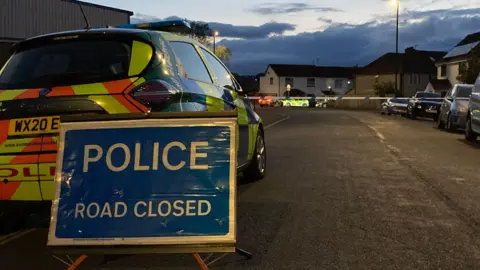 BBC A 'police - road closed' sign in the middle of the road, in front of a police car. A line of police tape can be seen across the road. 