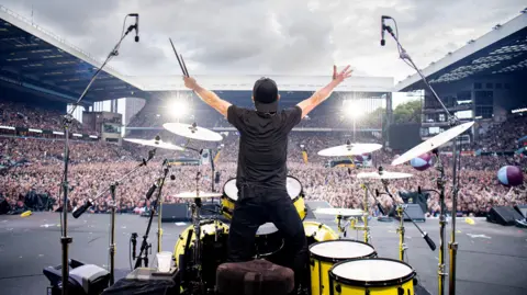 Ross Halfin A man stands behind a drumkit on a stage and holds his arms up to a large crowd. He is wearing all black and a black baseball cap that is on backwards.