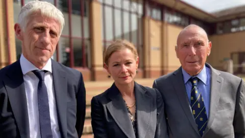 Briony Leyland/BBC A man in a black suit and tie, standing next to a woman in a dark suit and she is standing next to another man wearing a dark grey suit. They are standing in front of Surrey Coroner's Court.