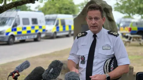 Stuart Woodward/BBC Ben-Julian Harrington wears a white shirt and black tie as he addresses the media while standing outdoors.
