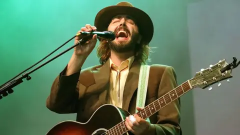 Getty Images Ben Schneider in a brown suit and hat holding a guitar and singing into a microphone on stage