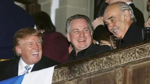 Getty Images Trump, McConnell and Sir Sean Connery enjoying a laugh on a balcony at an event at Tartan Week. They are all in dark business suits and we can see a bit of a Saltire in front of them.