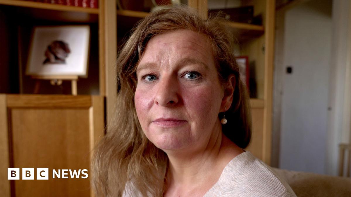 A headshot of Sarah, looking straight at the camera with a serious expression. She is wearing an oat coloured top and earrings with her hair loose. She is sitting in a living room, there are wooden shelves behind her.