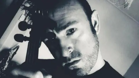 Ben Gunnery Ben Gunnery is looking directly at the camera. The image is black and white and he is holding a violin. He has dark stubble on his chin and short dark hair.