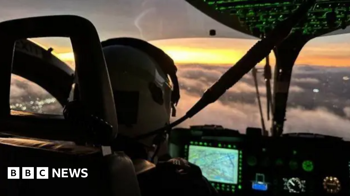 A close up of the inside of a police helicopter. The pilot is wearing a helmet and is sitting in front of a dashboard. The helicopter is high in the air above clouds.