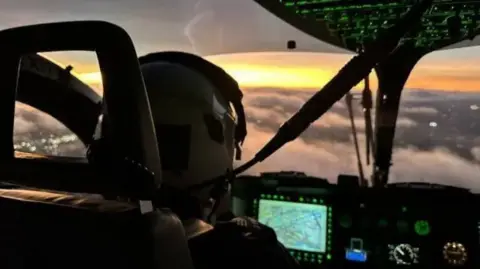 NPAS A close up of the inside of a police helicopter. The pilot is wearing a helmet and is sitting in front of a dashboard. The helicopter is high in the air above clouds.