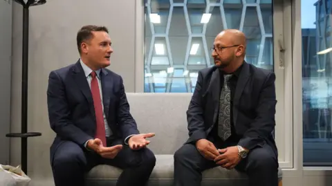 PA Media Two men sit next to each other on a grey seat and are facing each other in conversation. The man on the left wears a blue suit with red tie and is gesturing with his hands. The man on the right is wearing a blue suit and silver tie. He is wearing glasses and his hands are clasped in his lap.