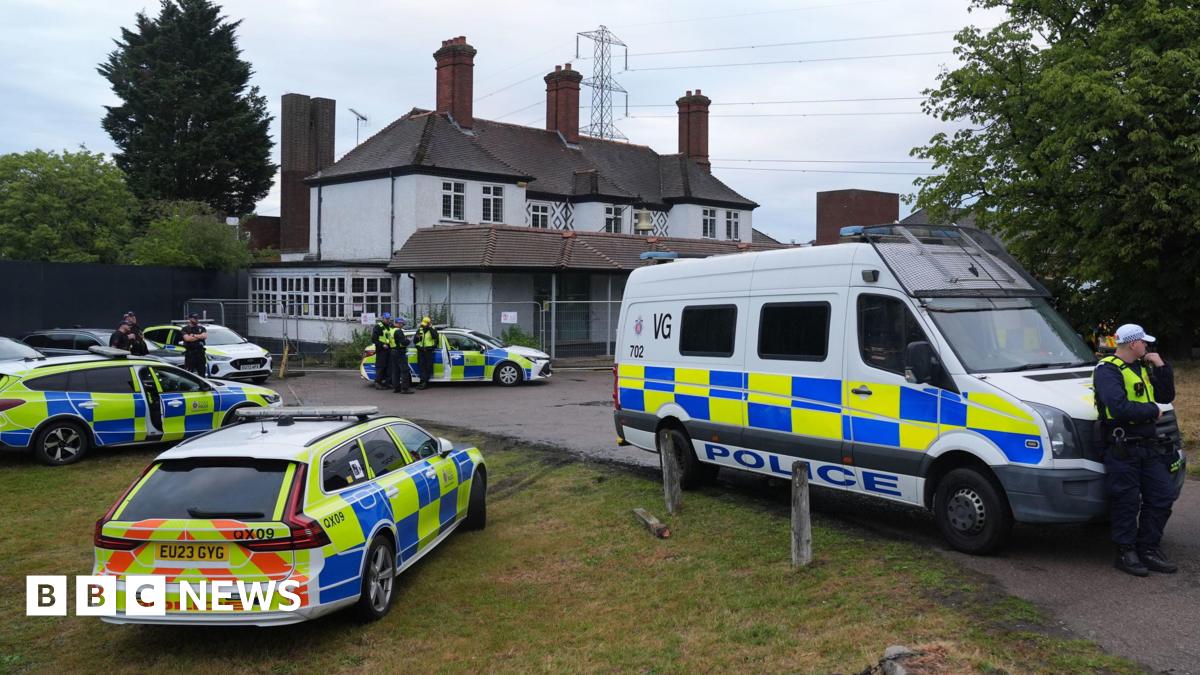 Multiple police cars and a van parked outside the grey hotel building, which is protected by fences. Several officers are standing around on watch.
