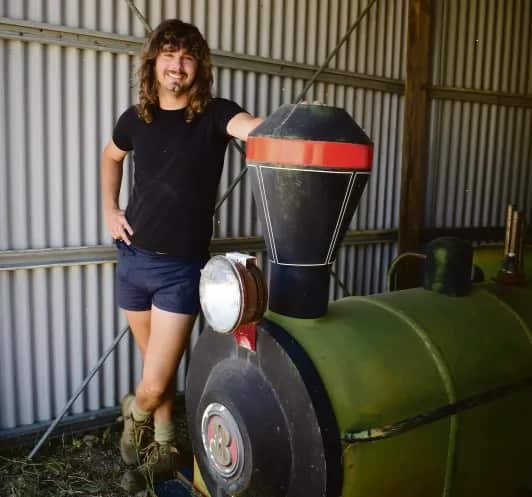 A young man with long, curly hair stands next to a miniature train.