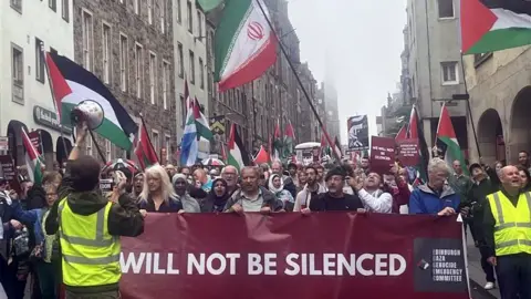 BBC A large protest on the Royal Mile in Edinburgh with several people marching behind a large banner. Some are waving flags of Palestine and Iran. One person has a blue and white Scottish flag.