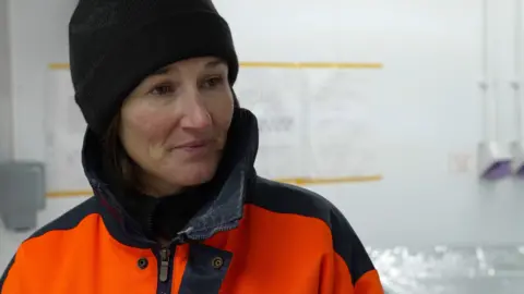 BBC News Liz Thomas stands inside a laboratory wearing an orange and black protective suit, and a hat. In the background there is a sign on the wall, and hand sanitiser dispensers.