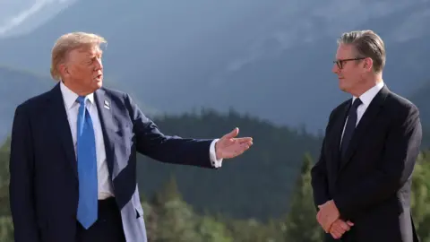Getty Images President Donald Trump gestures towards British Prime Minister Keir Starmer with his outstretched left hand. He is wearing a navy suit, white shirt and light blue tie. Sir Keir, who is standing with his arms folded is wearing a black suit, white shirt and navy tie. The leaders are standing in front of the Canadian Rockies. Lush green forests can be seen in the background