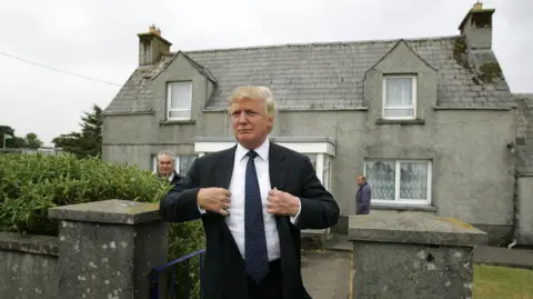 PA Media Donald Trump in dark blue suit, plain shite shirt and dark blue striped tie, standing outside a grey-harled house on a grey day. He is smiling and appears to be fixing his lapels.
