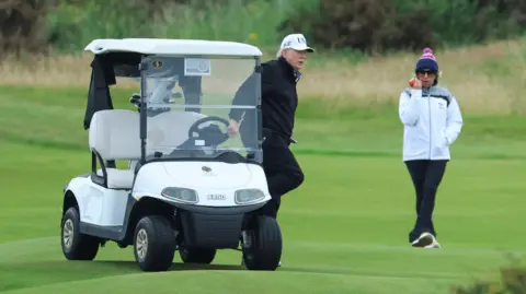 Reuters Donald Trump steps out of a golf buggy wearing dark-coloured waterproof golf clothes and a white cap. A woman wearing white waterproof jacket and 