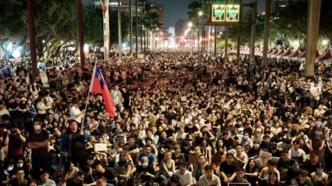 Getty Images A wide shot of thousands of protesters sitting on a boulevard in Taipei at night, holding up protest signs. In the foreground you can see a large Taiwan flag, while in the distance you can see the bright city lights of Taipei