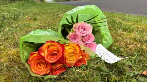 Pink and orange roses wrapped in a green plastic sleeves lie on the grass on a roadside with a white lined piece of paper which says  "We are heartbroken.  May they rest in peace. You are in our prayers."