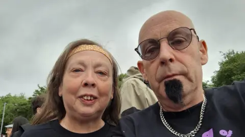 Simon Humphreys A man and a woman who are wearing Black Sabbath concert t-shirts looking at the camera. The man is bald and has a black goatee. The woman has brown hair and is wearing an orange headband 