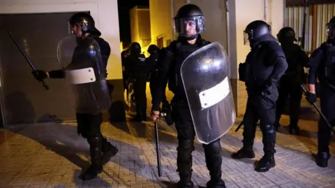 Reuters Riot police officers, seen in black uniform with riot shields, helmets and bats, stand guard at an alley way