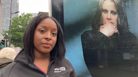 A woman with long black hair and wearing a black zip-up jacket which says "Westside Birmingham", standing in a Birmingham street. Behind her, to the right, is a poster of Ozzy Osbourne with his right hand up to his face.