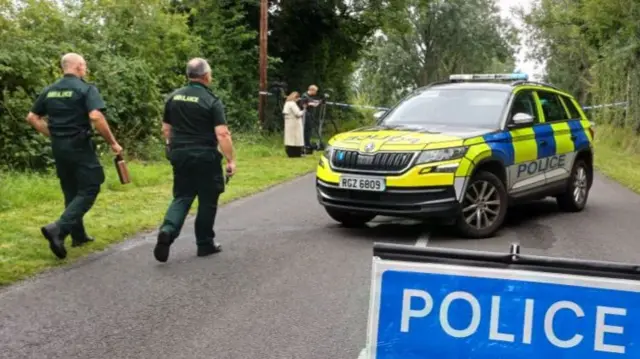Two male paramedics dressed in a green uniform walking towards blue and white tape. There is a yellow, blue and black police jeep with a blue 'police' sign.